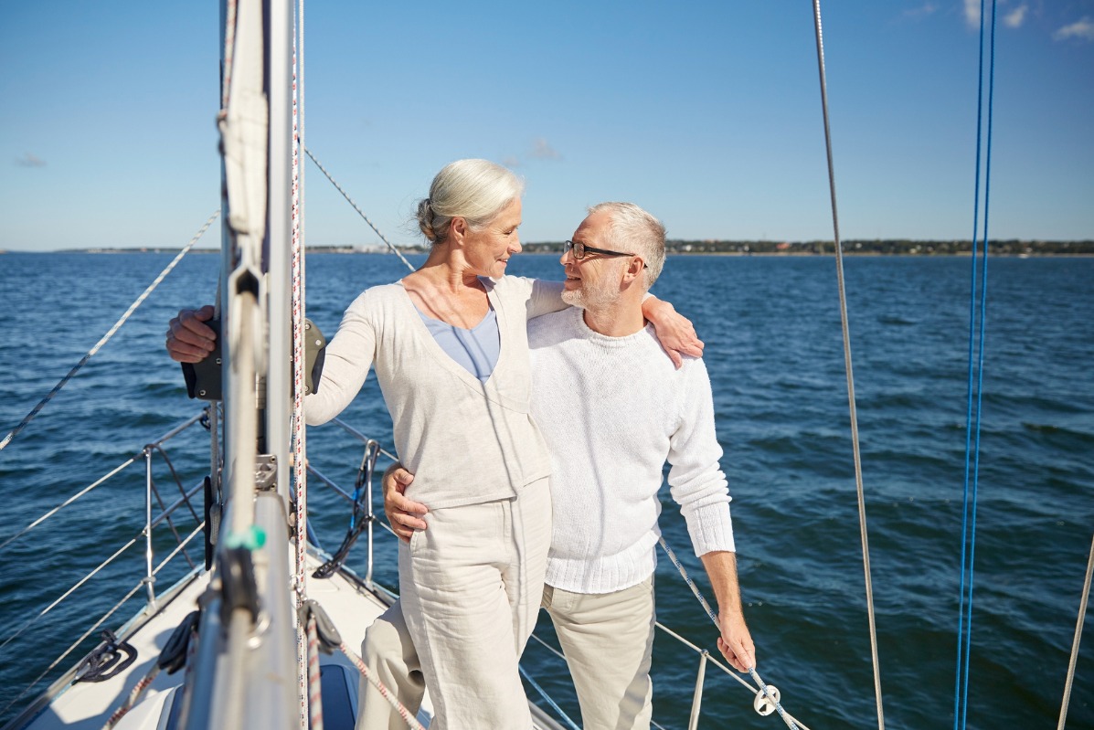 Para seniorów obejmuje się na pokładzie jachtu żaglowego, patrząc na siebie z uśmiechem, w tle błękitne morze i niebo.