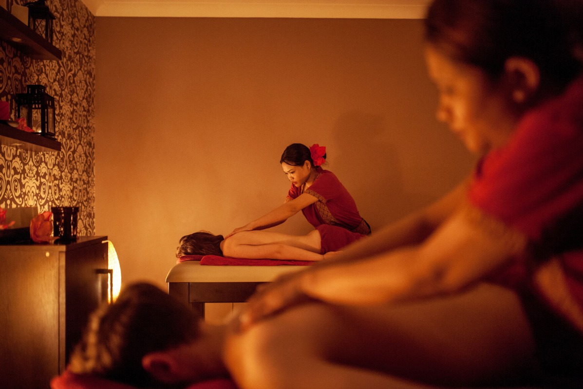 Masaż relaksacyjny w gabinecie: masażystka w czerwonej sukience z kwiatem we włosach masuje plecy klientki leżącej na stole do masażu, w tle wzorzysta tapeta i oświetlenie tworzące ciepłą atmosferę.