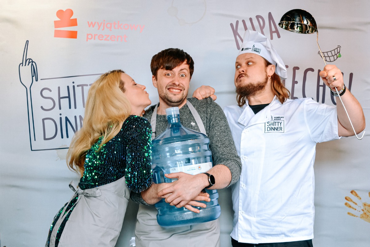 Trzy osoby w fartuchach pozują z dużym baniakiem wody i chochlą na tle ściany z napisem 'Wyjątkowy Prezent' i logo 'Shitty Dinner'.