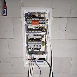 BARTMIX BARTOSZ PAHL - Rozdzielnica elektryczna w białej obudowie, zamontowana na ścianie z betonu komórkowego, z widocznymi przewodami i zabezpieczeniami, częściowo zakryta przez wystające kable. Na ścianie widoczne...