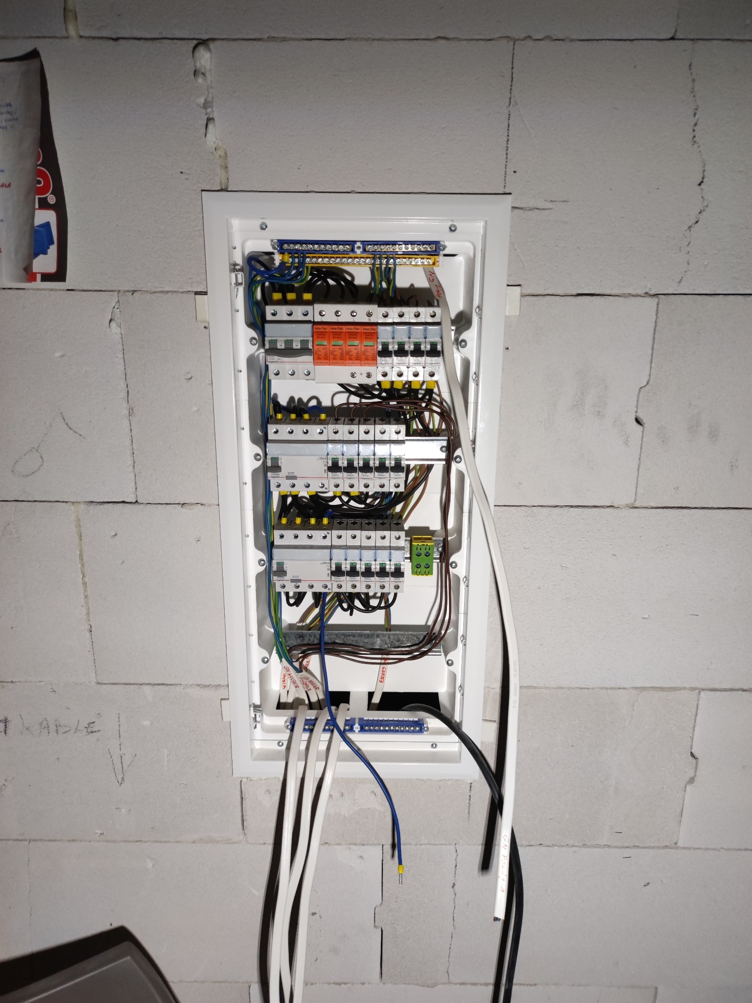 Rozdzielnica elektryczna w białej obudowie, zamontowana na ścianie z betonu komórkowego, z widocznymi przewodami i zabezpieczeniami, częściowo zakryta przez wystające kable. Na ścianie widoczne...