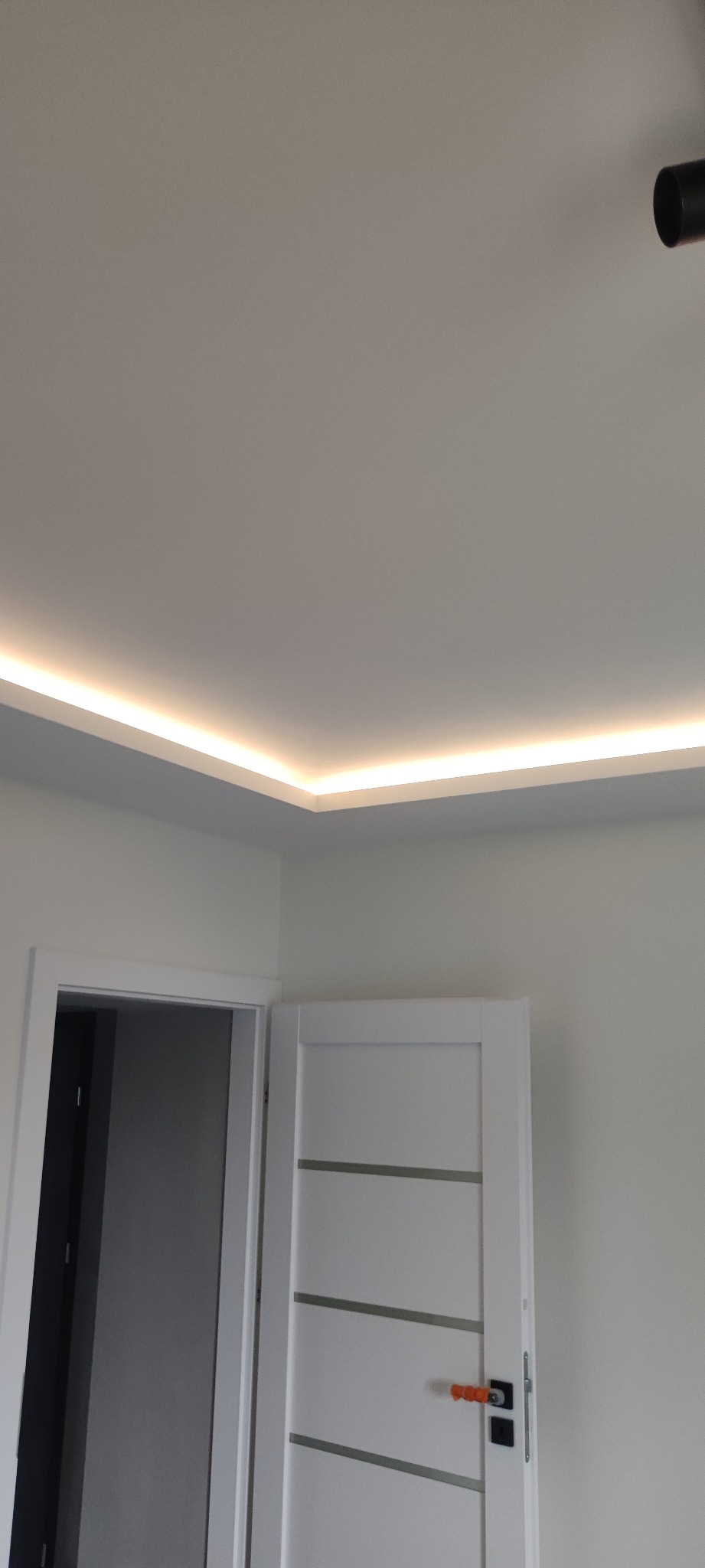 Nowoczesne oświetlenie LED w suficie podwieszanym, tworzące ciepłą linię światła wzdłuż krawędzi, z widocznymi białymi drzwiami z poziomymi wstawkami i fragmentem czarnego reflektora sufitowego.