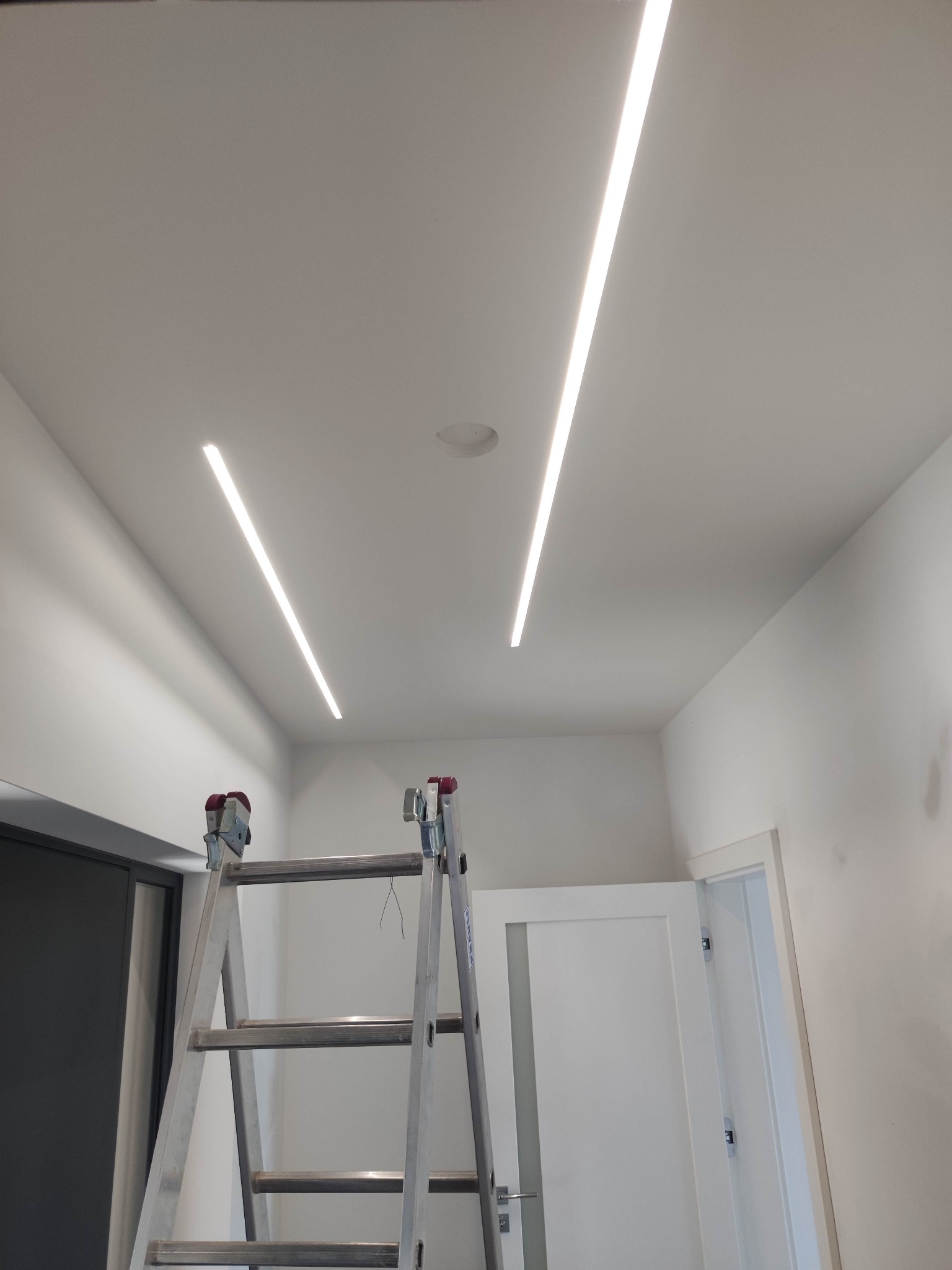 Montaż dwóch liniowych lamp LED w suficie podwieszanym, widoczna drabina aluminiowa z wystającymi przewodami.