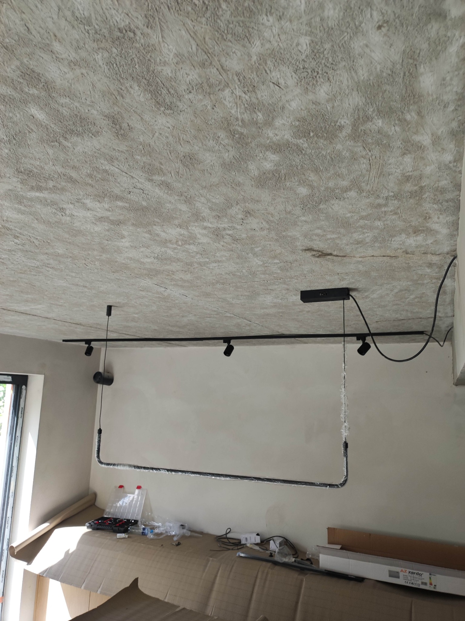 Szyna z reflektorami zamontowana na suficie w surowym stanie, widoczne narzędzia i opakowania po produktach na podłodze.