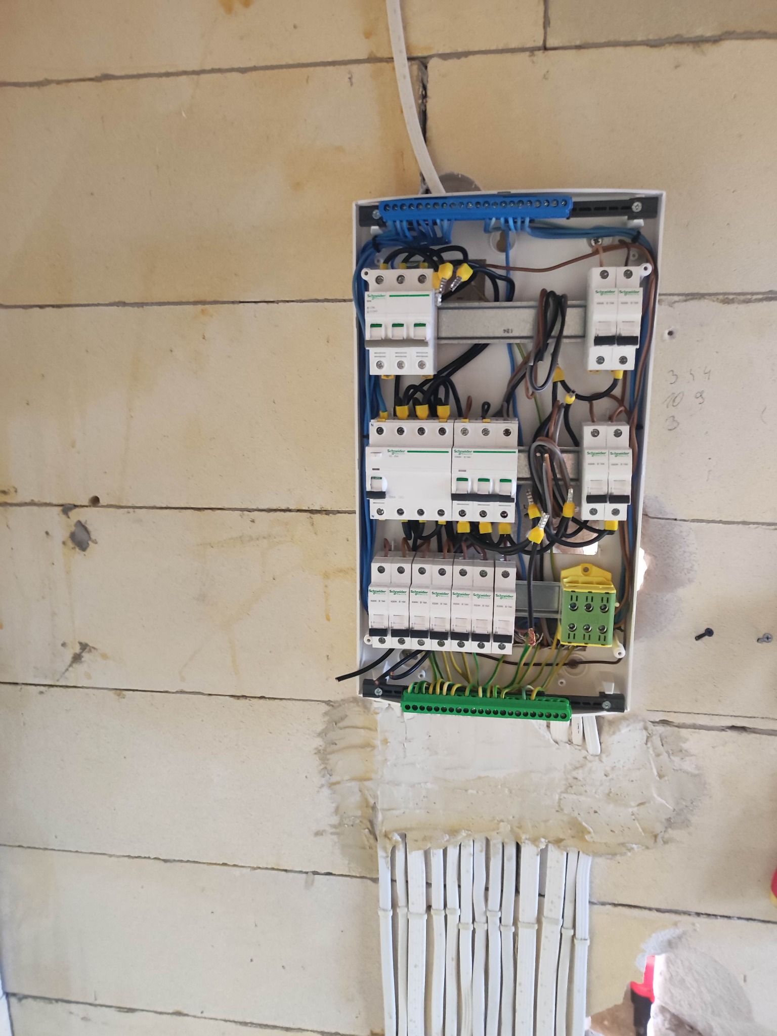 Rozdzielnica elektryczna z zainstalowanymi bezpiecznikami i przewodami, zamontowana na surowej ścianie z betonu komórkowego, z widocznymi przewodami w peszlach wchodzącymi do ściany.