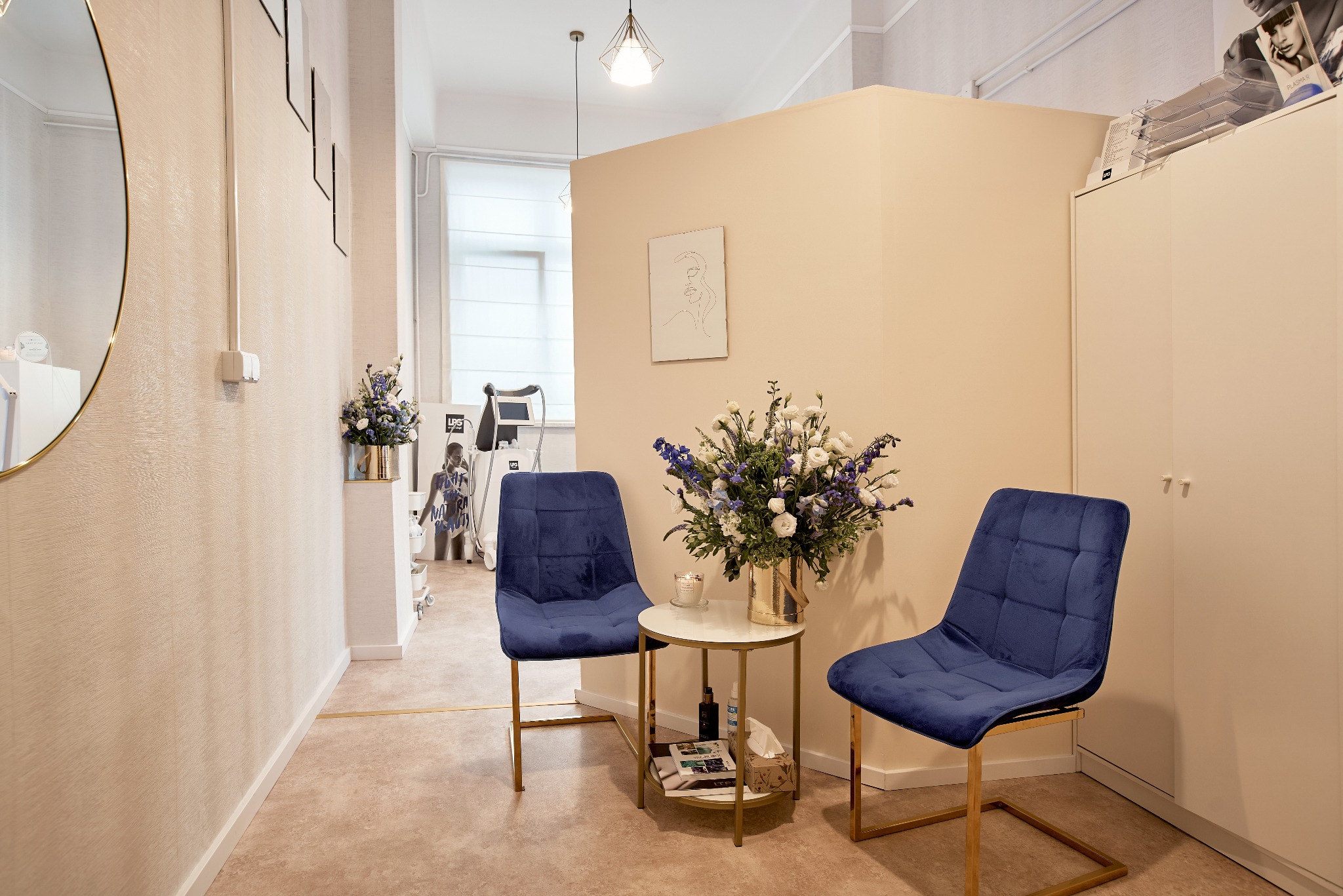 Jasne wnętrze poczekalni z dwoma granatowymi krzesłami i stolikiem z kwiatami, w tle widoczny sprzęt kosmetyczny i minimalistyczne dekoracje ścienne.