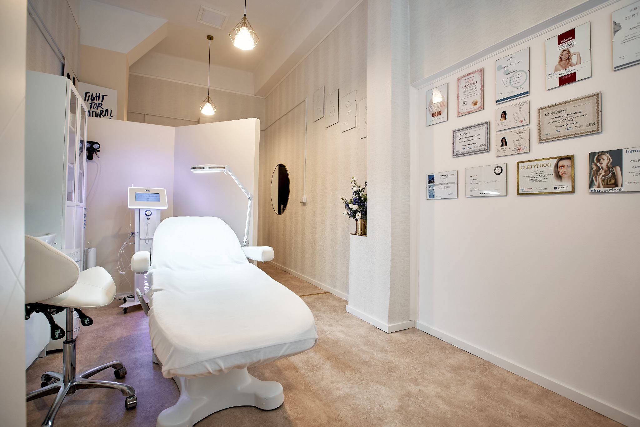 Jasne wnętrze gabinetu z białym fotelem kosmetycznym, lampą zabiegową i urządzeniem do zabiegów, ściany zdobią certyfikaty i dyplomy, a całość dopełniają dekoracje roślinne i designerskie lampy.