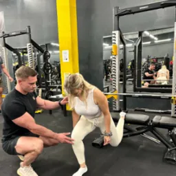 Trener asekuruje kobietę podczas przysiadu bułgarskiego na ławeczce w siłowni. W tle widoczne maszyny do ćwiczeń i lustra.