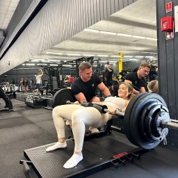 Trener personalny Wrocław Marcin Czarnecki - Kobieta ćwiczy unoszenie bioder ze sztangą, asystowana przez trenera w siłowni Evolution. Widoczne lustra i sprzęt do ćwiczeń. Uśmiechnięta twarz kobiety.