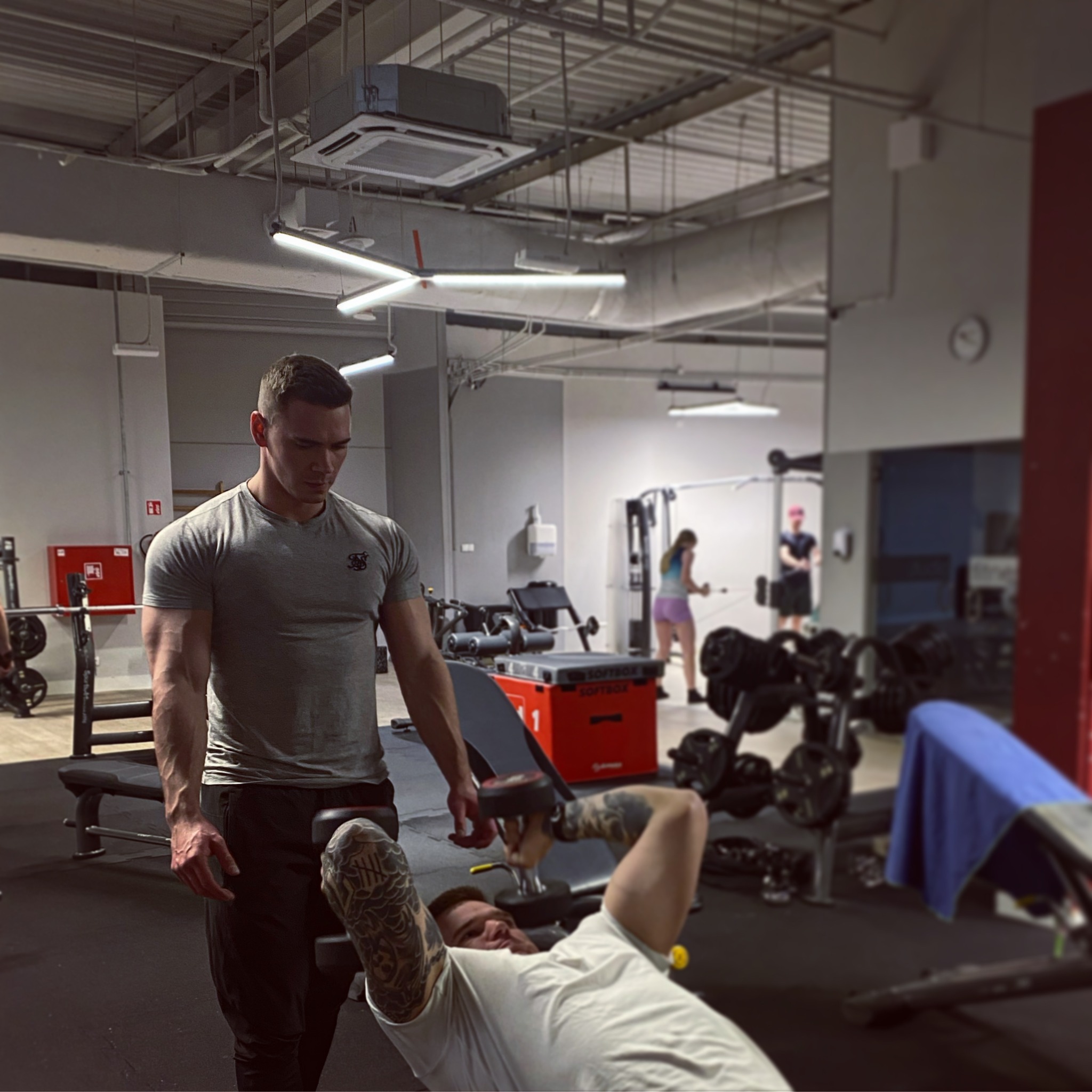 Mężczyzna z tatuażami na ramionach ćwiczy wyciskanie hantli leżąc na ławce, nadzorowany przez trenera w szarej koszulce w siłowni.