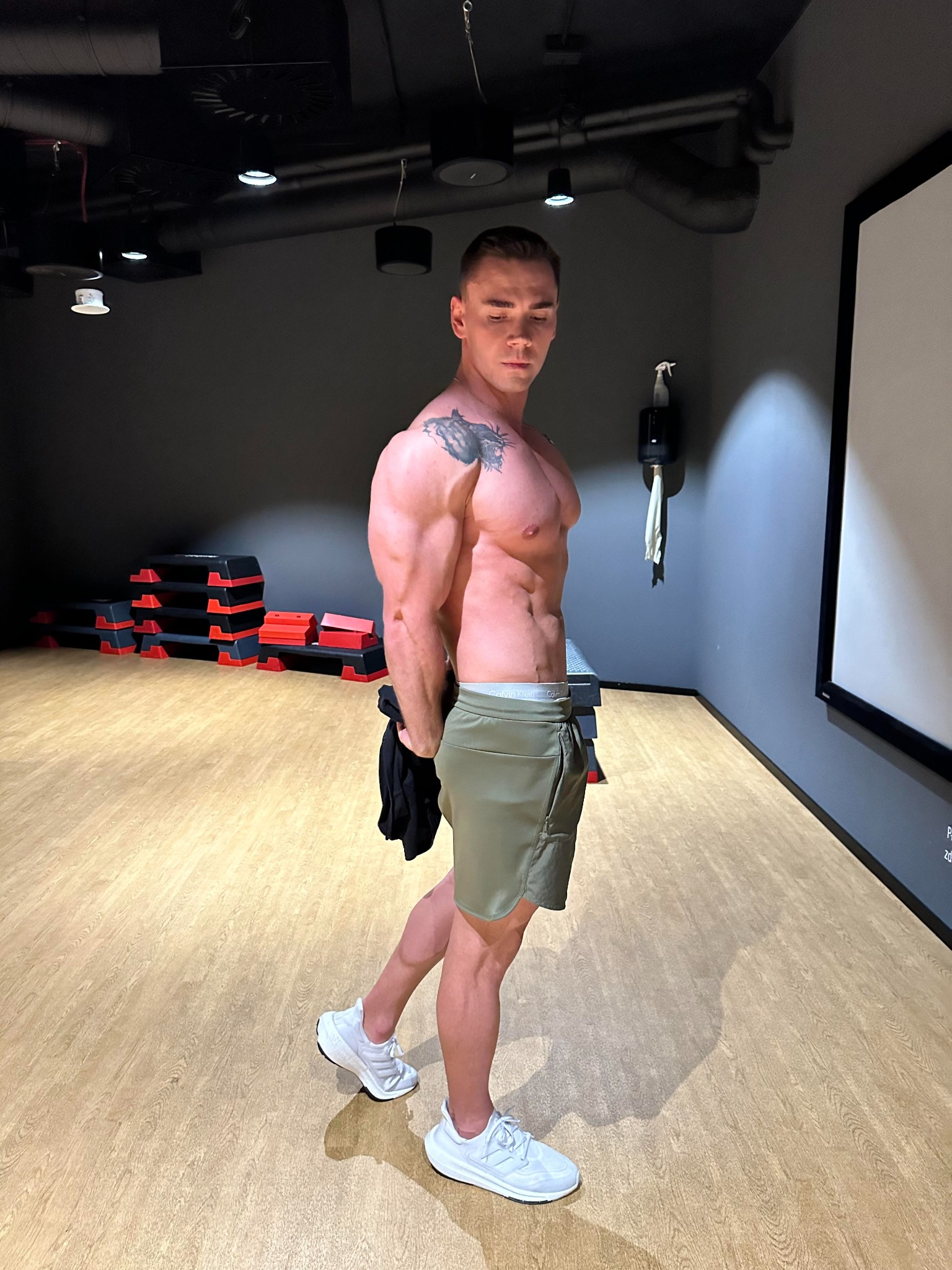 Mężczyzna prezentuje wyrzeźbioną sylwetkę w siłowni, widoczne stepy do ćwiczeń w tle, tatuaż na ramieniu, ubrany w szorty khaki i białe sportowe buty.