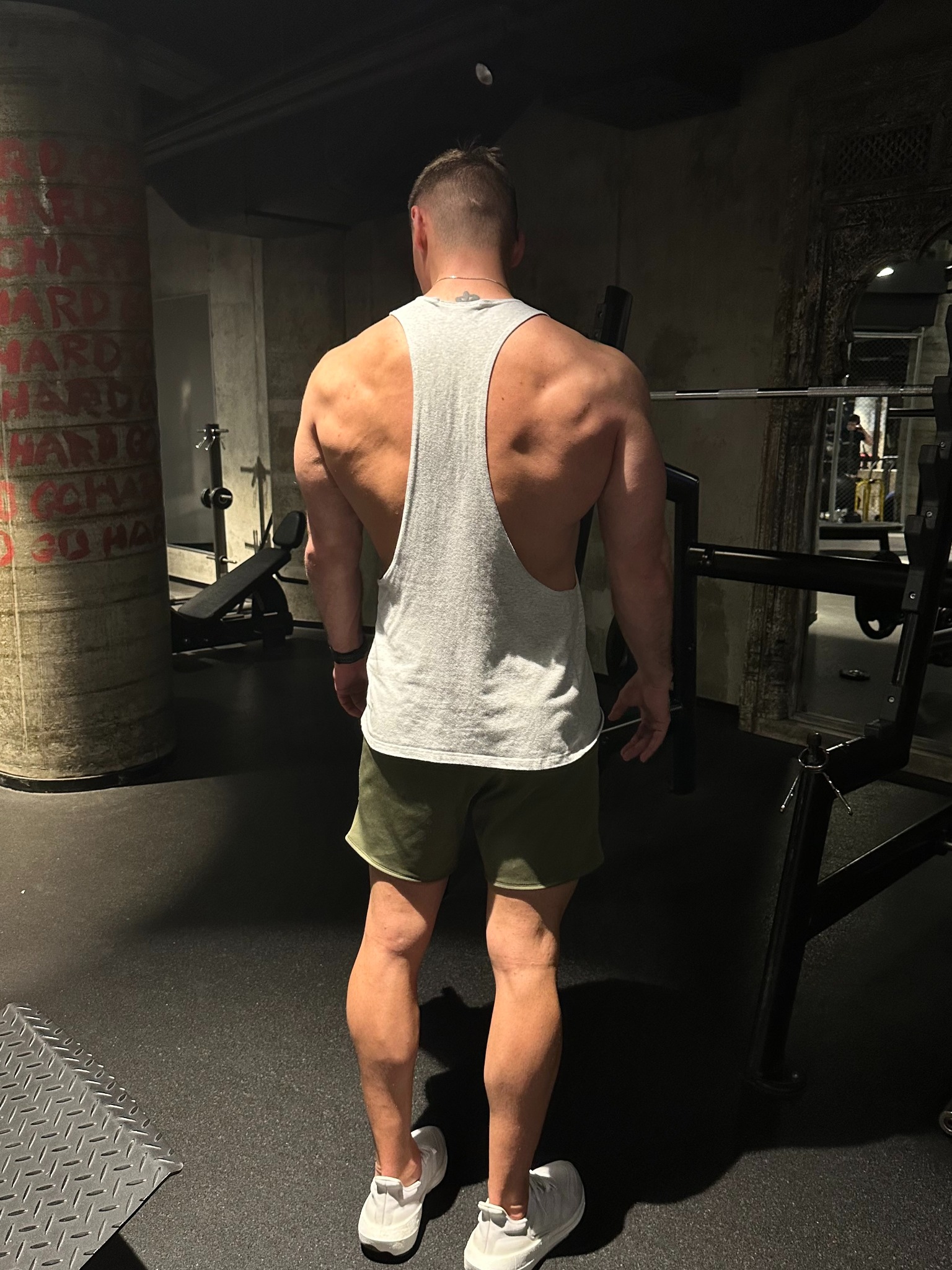 Mężczyzna w szarej koszulce bez rękawów i zielonych spodenkach stoi tyłem w ciemnej siłowni, prezentując muskularne plecy i ramiona, widoczne sprzęty do ćwiczeń w tle.