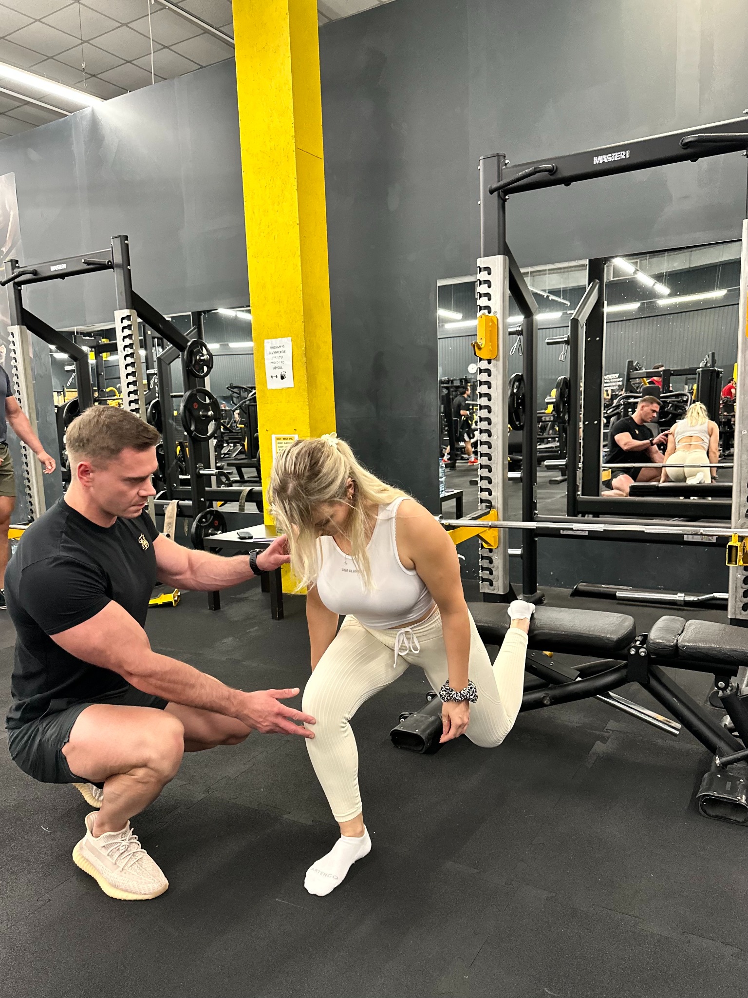 Trener asekuruje kobietę podczas przysiadu bułgarskiego na ławeczce w siłowni. W tle widoczne maszyny do ćwiczeń i lustra.