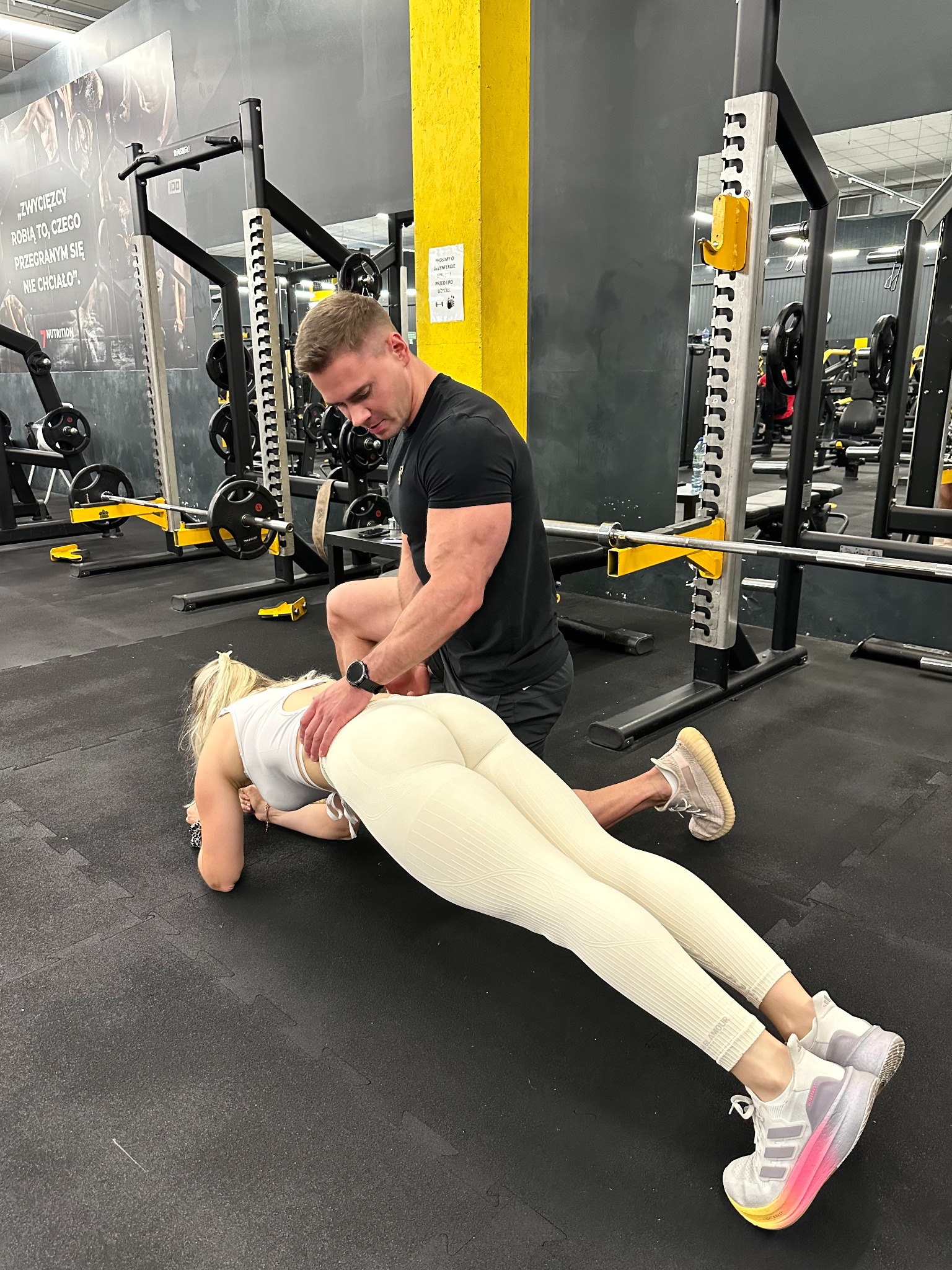 Trener personalny koryguje postawę kobiety podczas ćwiczenia plank w siłowni. Kobieta w białym stroju, trener w czarnym. W tle sprzęt do ćwiczeń siłowych.