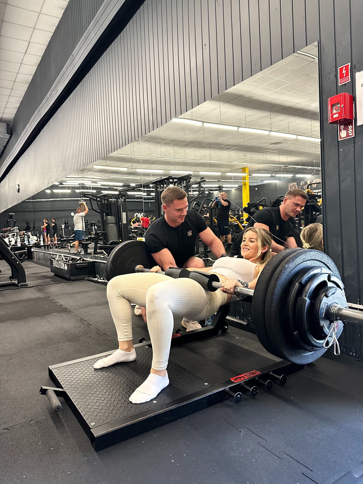 Kobieta ćwiczy unoszenie bioder ze sztangą, asystowana przez trenera w siłowni Evolution. Widoczne lustra i sprzęt do ćwiczeń. Uśmiechnięta twarz kobiety.