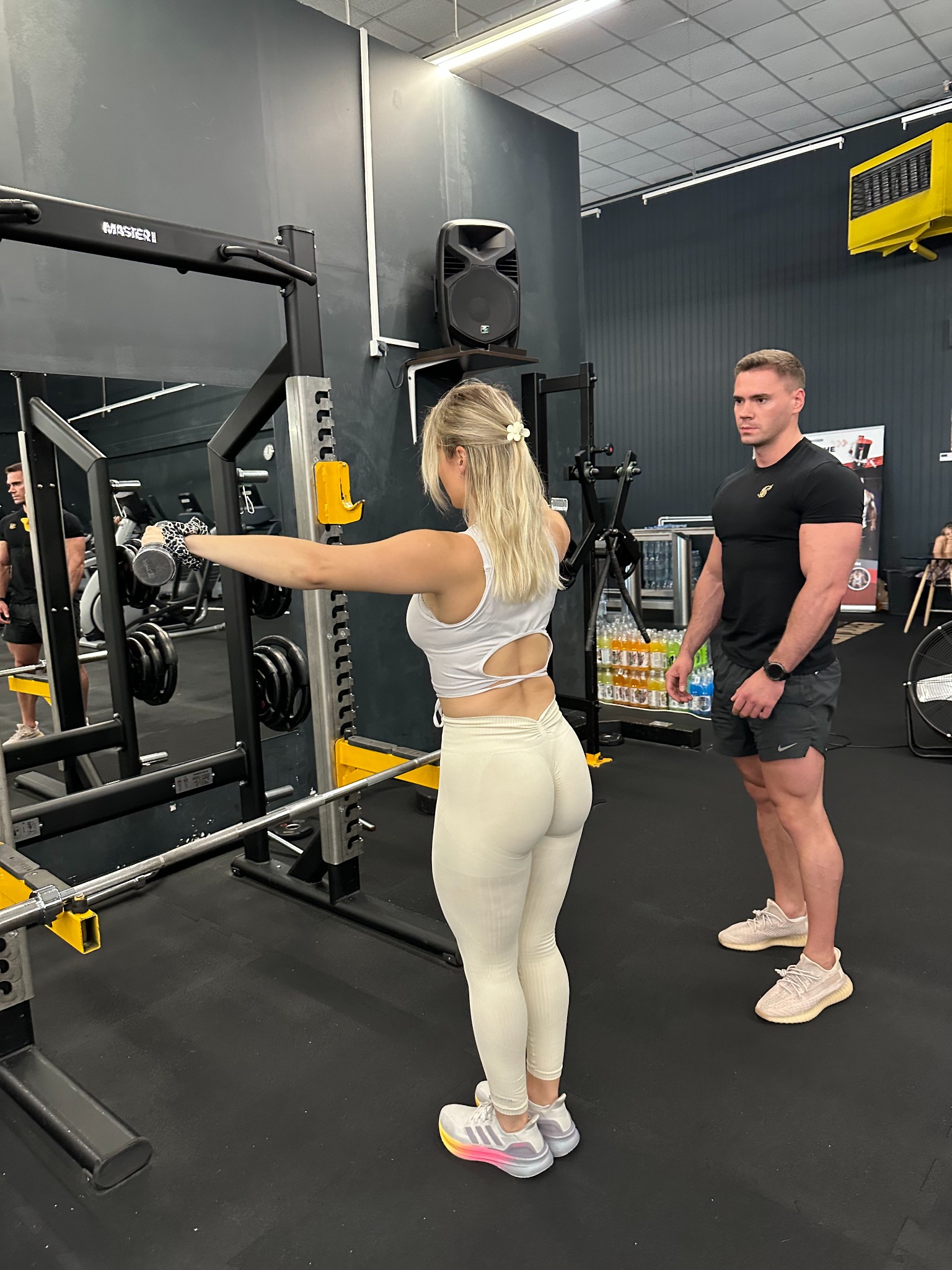 Kobieta ćwiczy unoszenie hantli pod okiem trenera w siłowni. Widoczny sprzęt do ćwiczeń, lustro i butelki z wodą w tle. Kontrastowe, czarno-białe wnętrze.