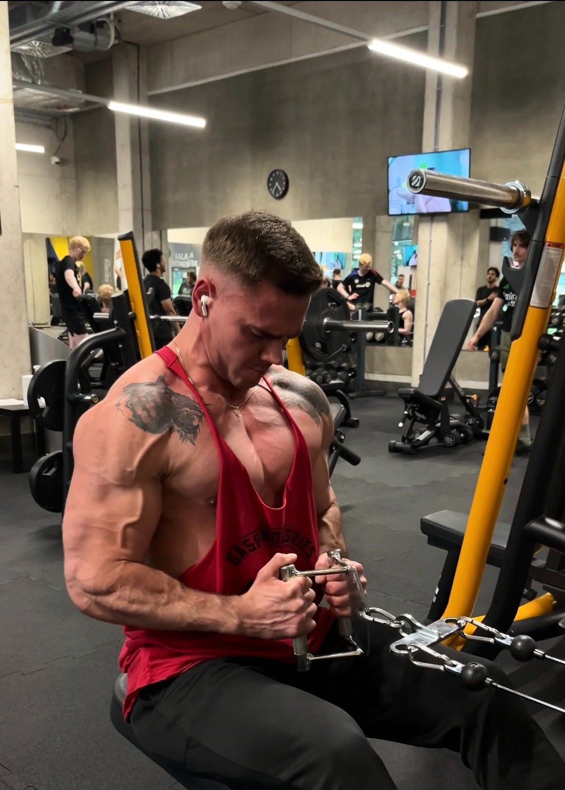Mężczyzna z tatuażem na ramieniu ćwiczy na maszynie w siłowni we Wrocławiu. Widoczne napięte mięśnie i czerwona koszulka bez rękawów.