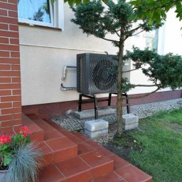 SUN ENERGY INDUSTRIES PATRYK GIZLER - Zamontowana pompa ciepła powietrze-woda na zewnątrz budynku, obok wejścia z czerwonymi schodami i ozdobnym drzewkiem. Widoczne rury i podstawa pompy.