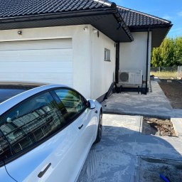 SUN ENERGY INDUSTRIES PATRYK GIZLER - Biały budynek z garażem i zamontowaną pompą ciepła Panasonic. Obok zaparkowany biały samochód Tesla. Widoczne prace brukarskie.