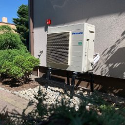 SUN ENERGY INDUSTRIES PATRYK GIZLER - Kompaktowa pompa ciepła Panasonic R32 na czarnym stelażu, zamontowana przy ścianie budynku, otoczona zielenią i kamieniami ozdobnymi. Słoneczny dzień.