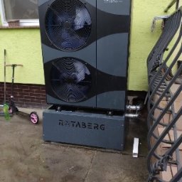 SUN ENERGY INDUSTRIES PATRYK GIZLER - Szara pompa ciepła Rotaberg z dwoma wentylatorami, zamontowana na zewnątrz budynku z żółtą elewacją, widoczna z poziomu gruntu, obok barierka i hulajnoga.