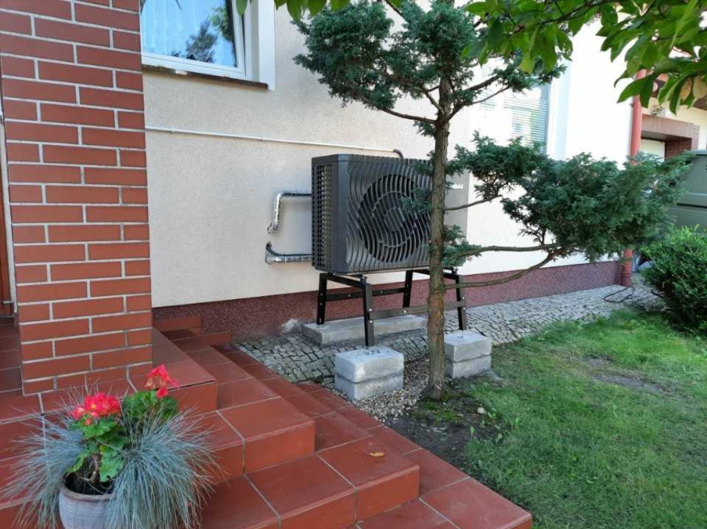 Zamontowana pompa ciepła powietrze-woda na zewnątrz budynku, obok wejścia z czerwonymi schodami i ozdobnym drzewkiem. Widoczne rury i podstawa pompy.