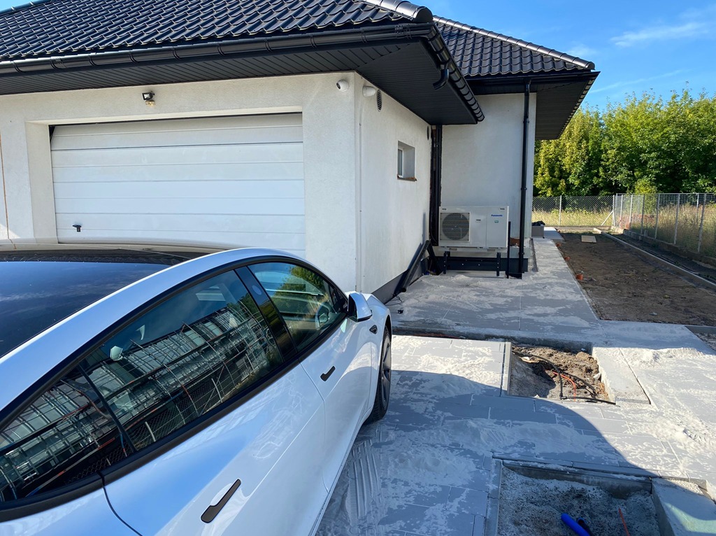 Biały budynek z garażem i zamontowaną pompą ciepła Panasonic. Obok zaparkowany biały samochód Tesla. Widoczne prace brukarskie.