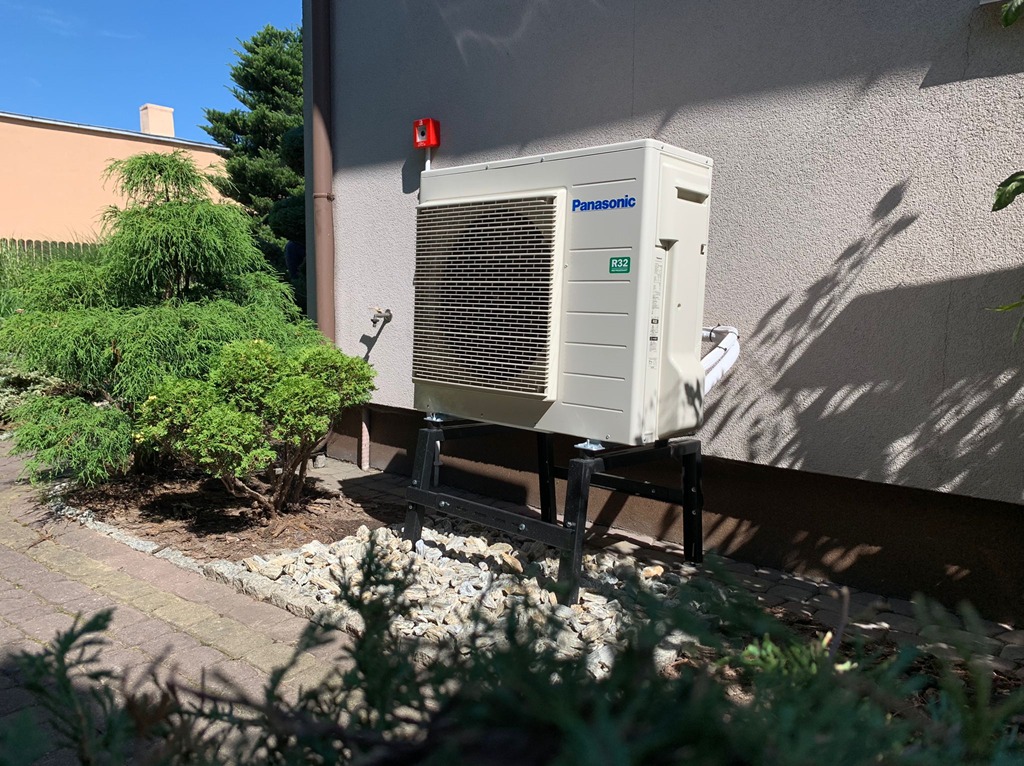 Kompaktowa pompa ciepła Panasonic R32 na czarnym stelażu, zamontowana przy ścianie budynku, otoczona zielenią i kamieniami ozdobnymi. Słoneczny dzień.