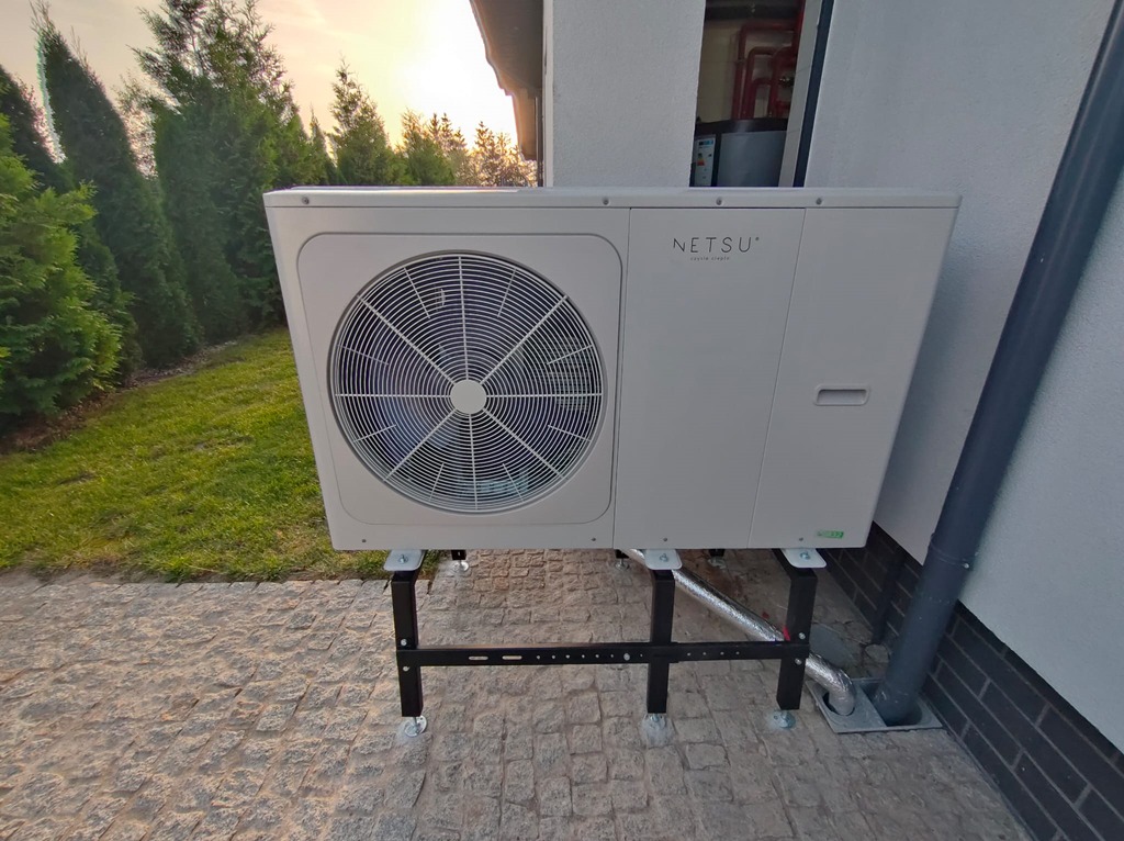 Biała pompa ciepła Netsu na czarnym stelażu, z widocznym wentylatorem, zainstalowana na zewnątrz budynku z zielonym trawnikiem i tujami w tle.