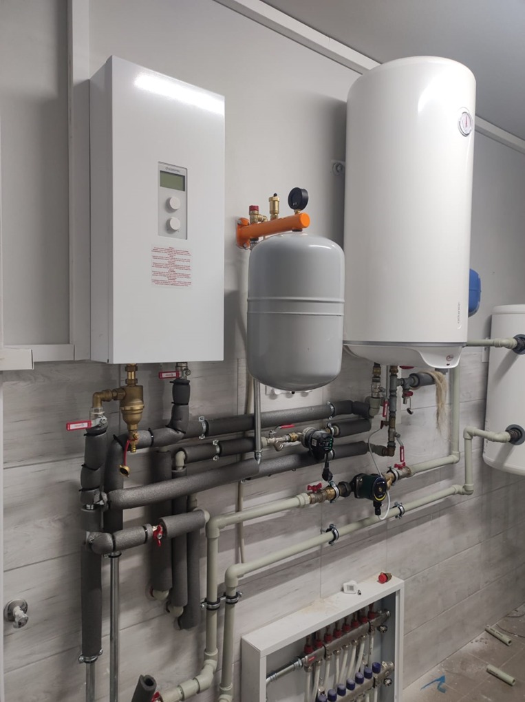 Biały piec grzewczy, zbiornik buforowy i rozdzielacz podłogówki na tle ściany z panelami imitującymi drewno. Widoczne liczne rury i zawory.