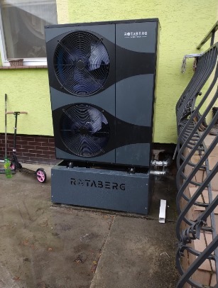 Szara pompa ciepła Rotaberg z dwoma wentylatorami, zamontowana na zewnątrz budynku z żółtą elewacją, widoczna z poziomu gruntu, obok barierka i hulajnoga.