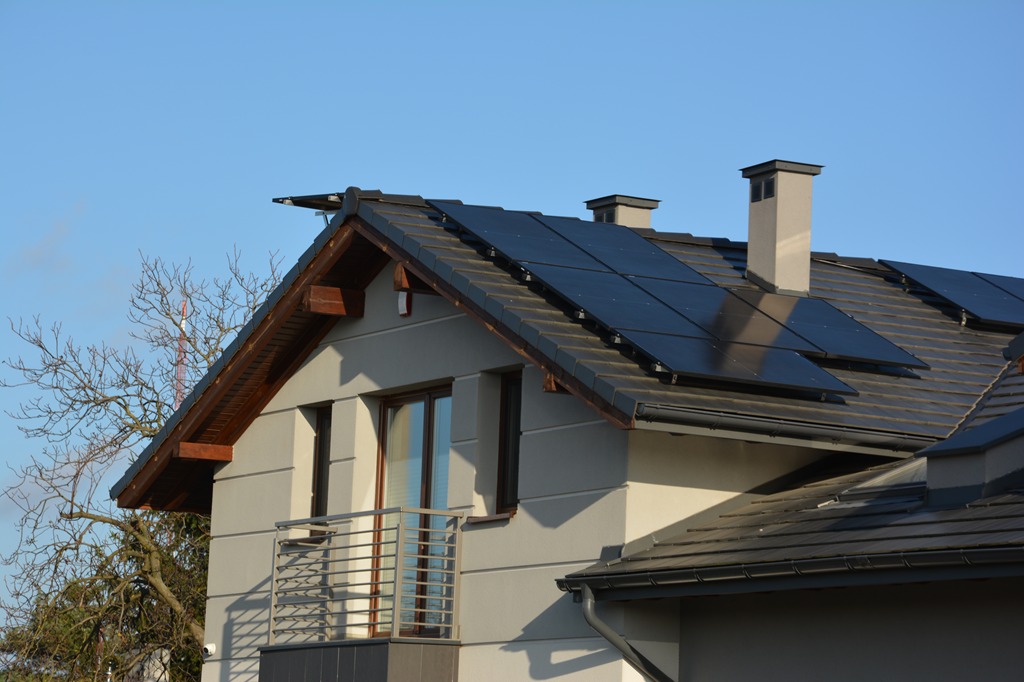 Częściowy widok domu z panelami słonecznymi na dachu z ciemnej dachówki, komin i balkon z metalową barierką. Błękitne niebo w tle.