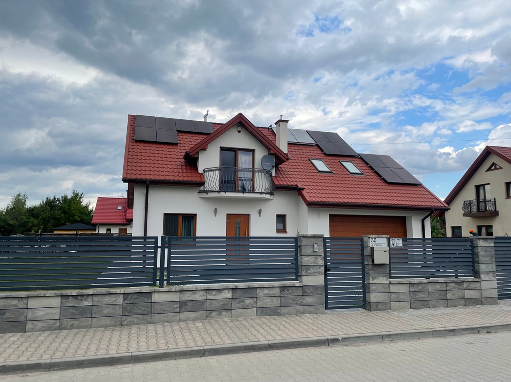 Dom jednorodzinny z panelami słonecznymi na czerwonym dachu, widoczny zza szarego ogrodzenia. Na elewacji balkon z czarną balustradą i satelitą.