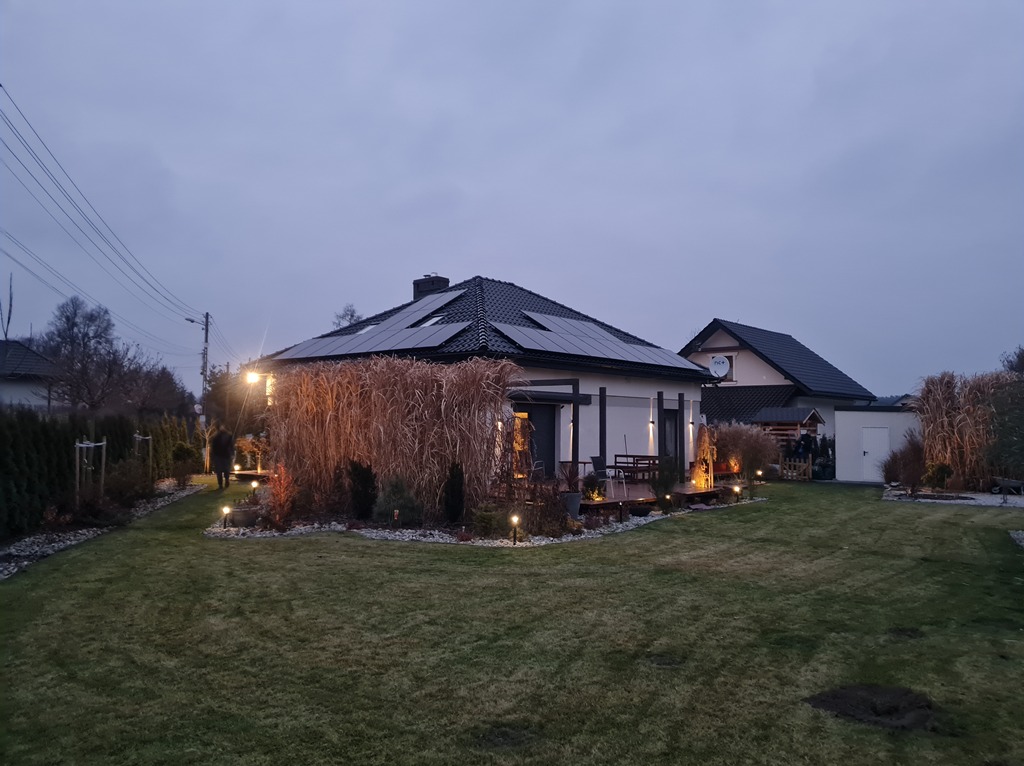 Dom jednorodzinny z panelami słonecznymi na dachu, oświetlony ogród z ozdobnymi trawami, widok wieczorny.