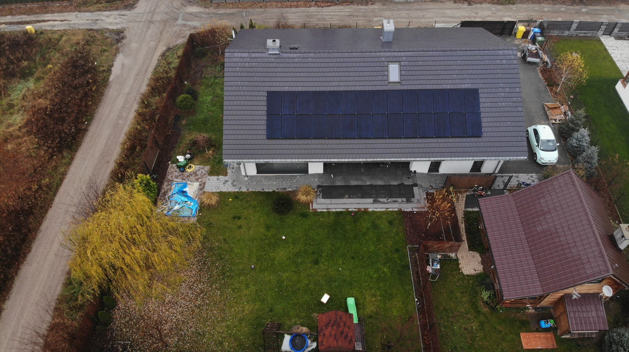 Widok z drona na dom z panelami słonecznymi na szarym dachu. Obok domu ogród, ścieżka i budynek gospodarczy z brązowym dachem.