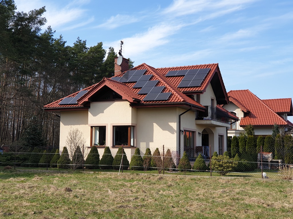 Dom jednorodzinny z panelami słonecznymi na czerwonym dachu, widok z ogrodu. Antena satelitarna na kominie. W tle drzewa i błękitne niebo.