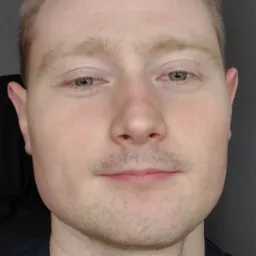 Portret mężczyzny z bliska, z lekkim zarostem i zaczerwienieniem na szyi, na tle ciemnej ściany.