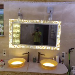 Krzysztof Karna - Oświetlona mozaiką łazienka z dwoma żółtymi umywalkami, ciemnymi bateriami i dużym lustrem, odbijającym postać fotografa.