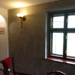 Krzysztof Karna - Ściana po malowaniu dekoracyjnym w odcieniach szarości, oświetlona kinkietem z ciepłym światłem, okno z drewnianą ramą, fragment krzesła z czerwoną czapką Mikołaja i taśma malarska na stole.