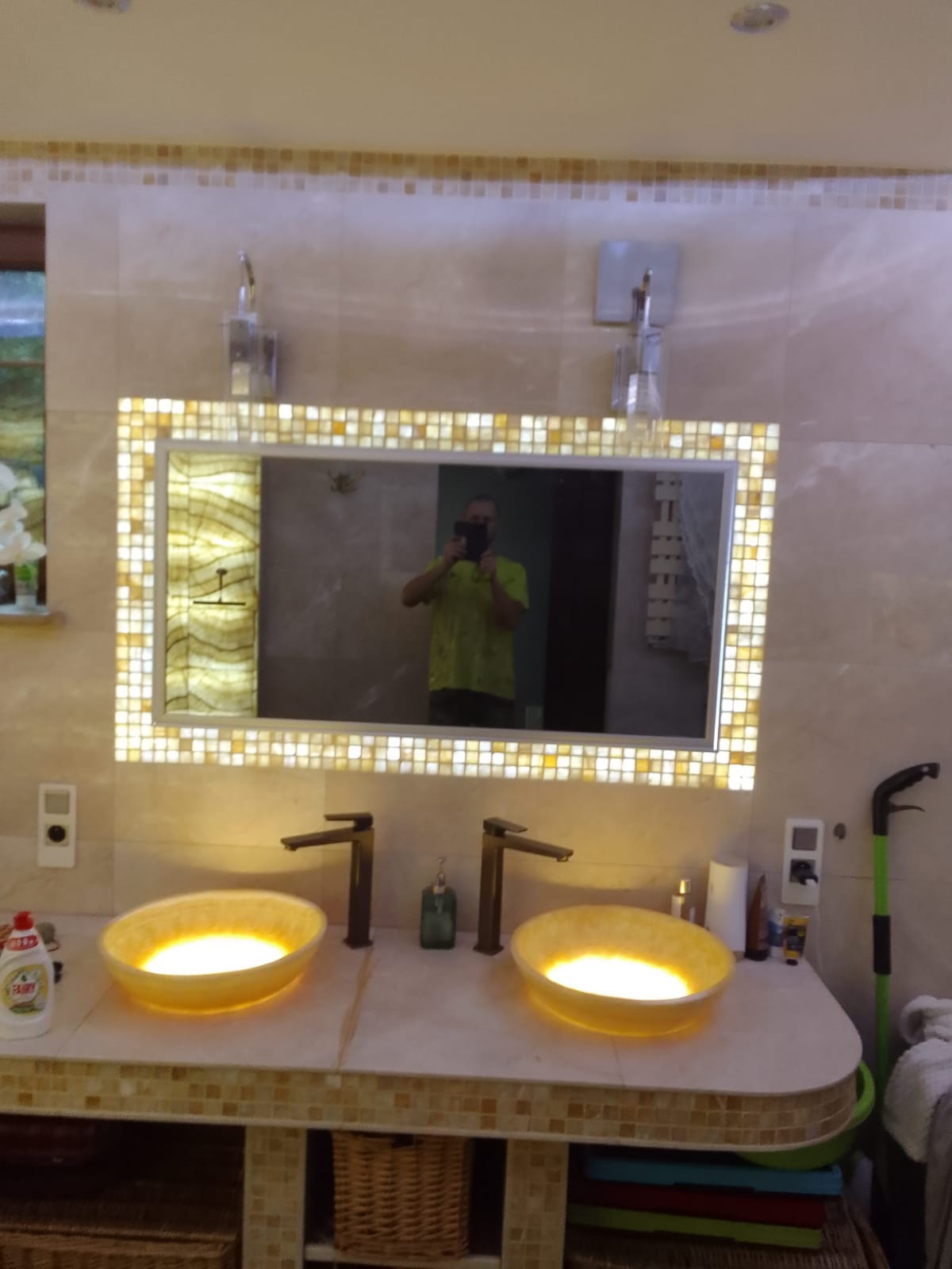 Oświetlona mozaiką łazienka z dwoma żółtymi umywalkami, ciemnymi bateriami i dużym lustrem, odbijającym postać fotografa.
