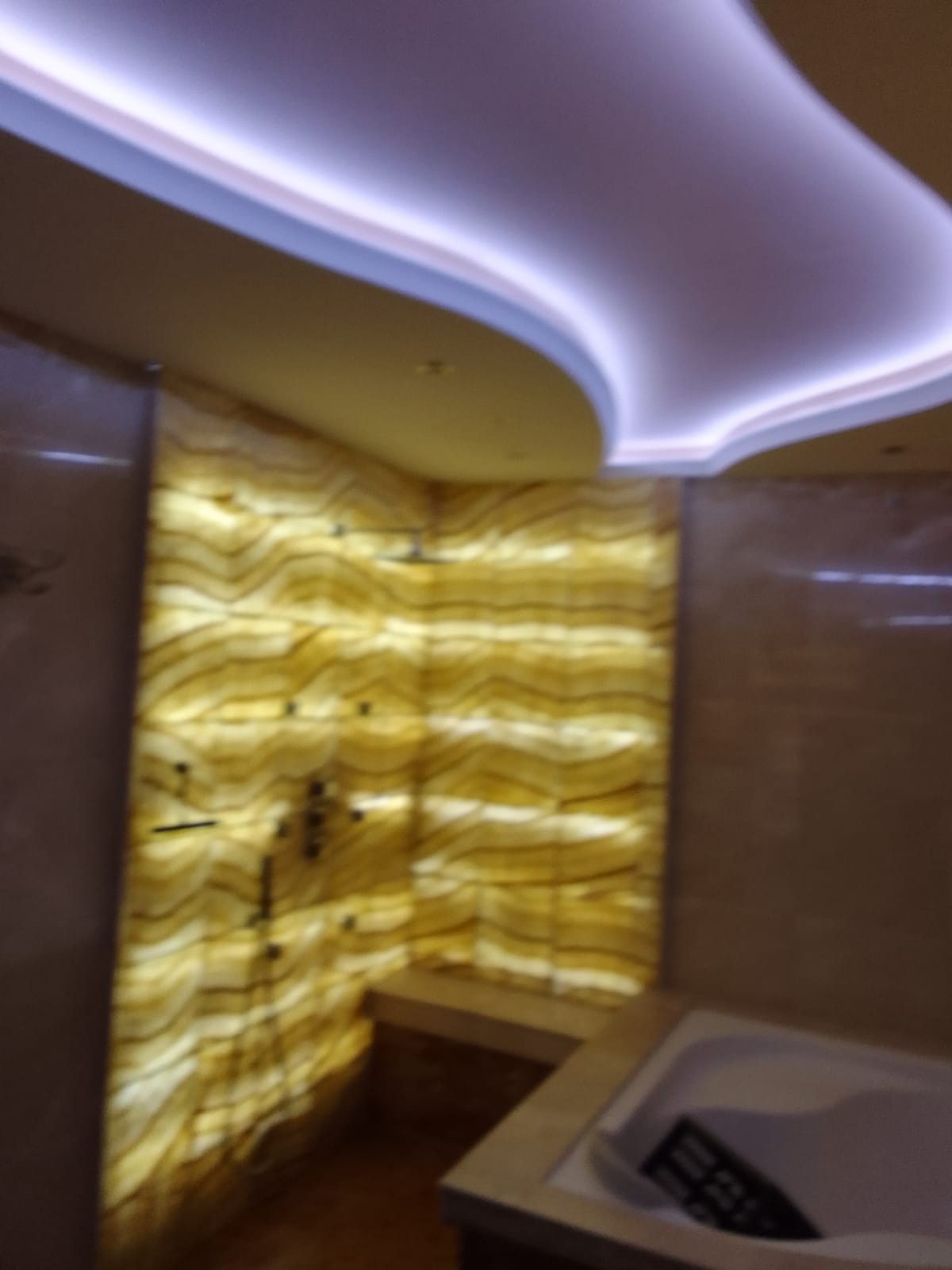 Nowoczesna łazienka z podświetlaną ścianą z onyksu, widoczny fragment wanny i dekoracyjne oświetlenie sufitowe.