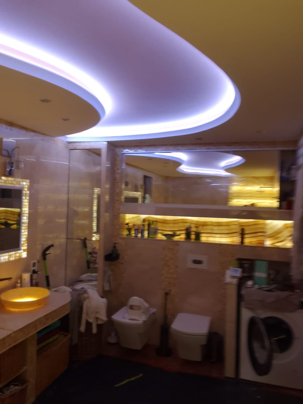 Nowoczesna łazienka z podwieszanym sufitem o nieregularnym kształcie z oświetleniem LED, marmurkowymi płytkami i podświetlaną półką z kosmetykami, widok na umywalkę z żółtą misą i pralkę.