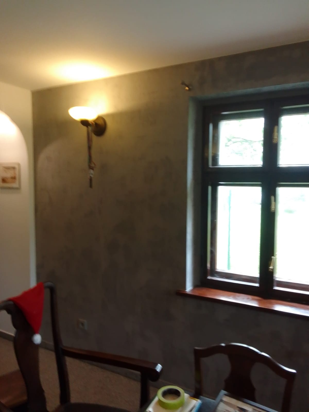 Ściana po malowaniu dekoracyjnym w odcieniach szarości, oświetlona kinkietem z ciepłym światłem, okno z drewnianą ramą, fragment krzesła z czerwoną czapką Mikołaja i taśma malarska na stole.
