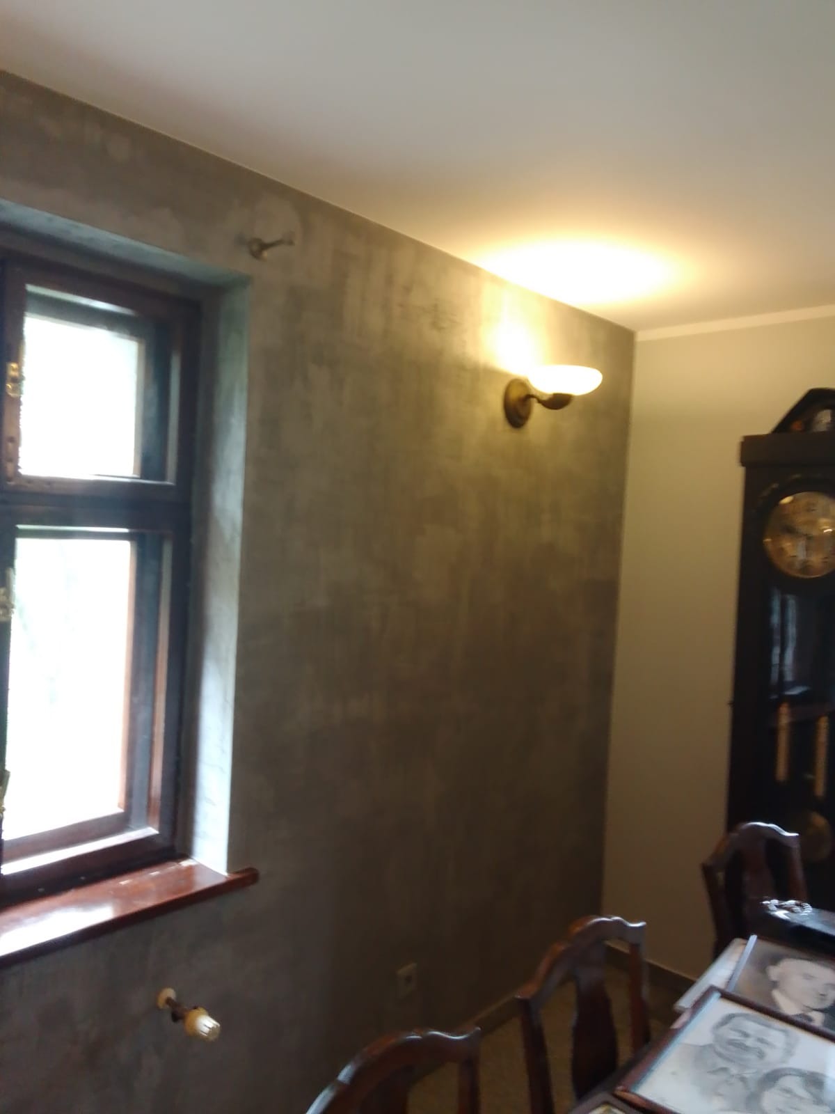 Ściana w pokoju po nałożeniu dekoracyjnej, szarej farby o nieregularnej fakturze, widoczne drewniane okno z parapetem, kinkiet na ścianie i fragment starego zegara.