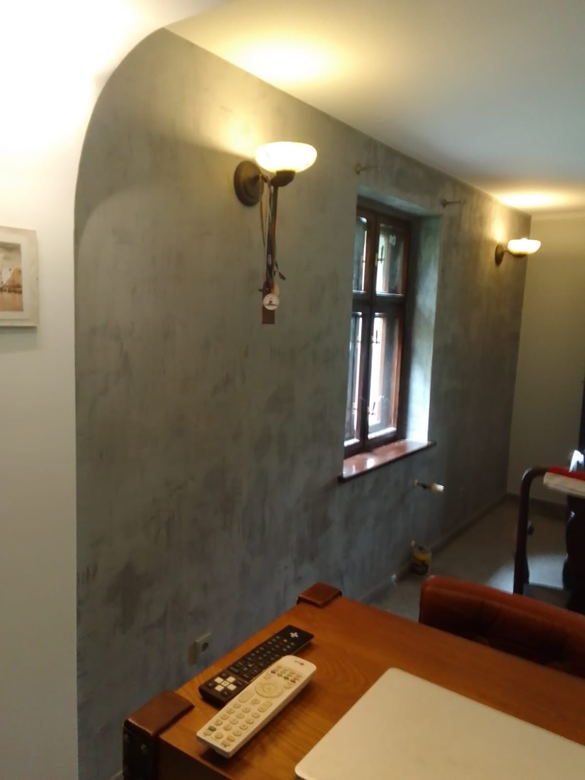 Ściana w pokoju z efektem dekoracyjnym, widoczne lampy ścienne z okrągłymi kloszami, drewniana rama okienna, dwa piloty leżące na drewnianym stole, oraz fragment laptopa.