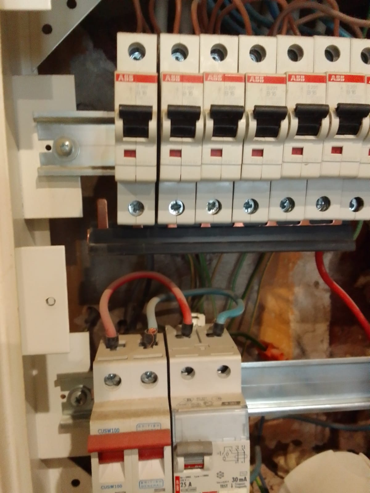 Szczegółowe ujęcie tablicy rozdzielczej z zamontowanymi bezpiecznikami ABB i przewodami elektrycznymi w kolorach czerwonym, zielonym i niebieskim, widoczne ślady po wierceniu w ścianie za tablicą.