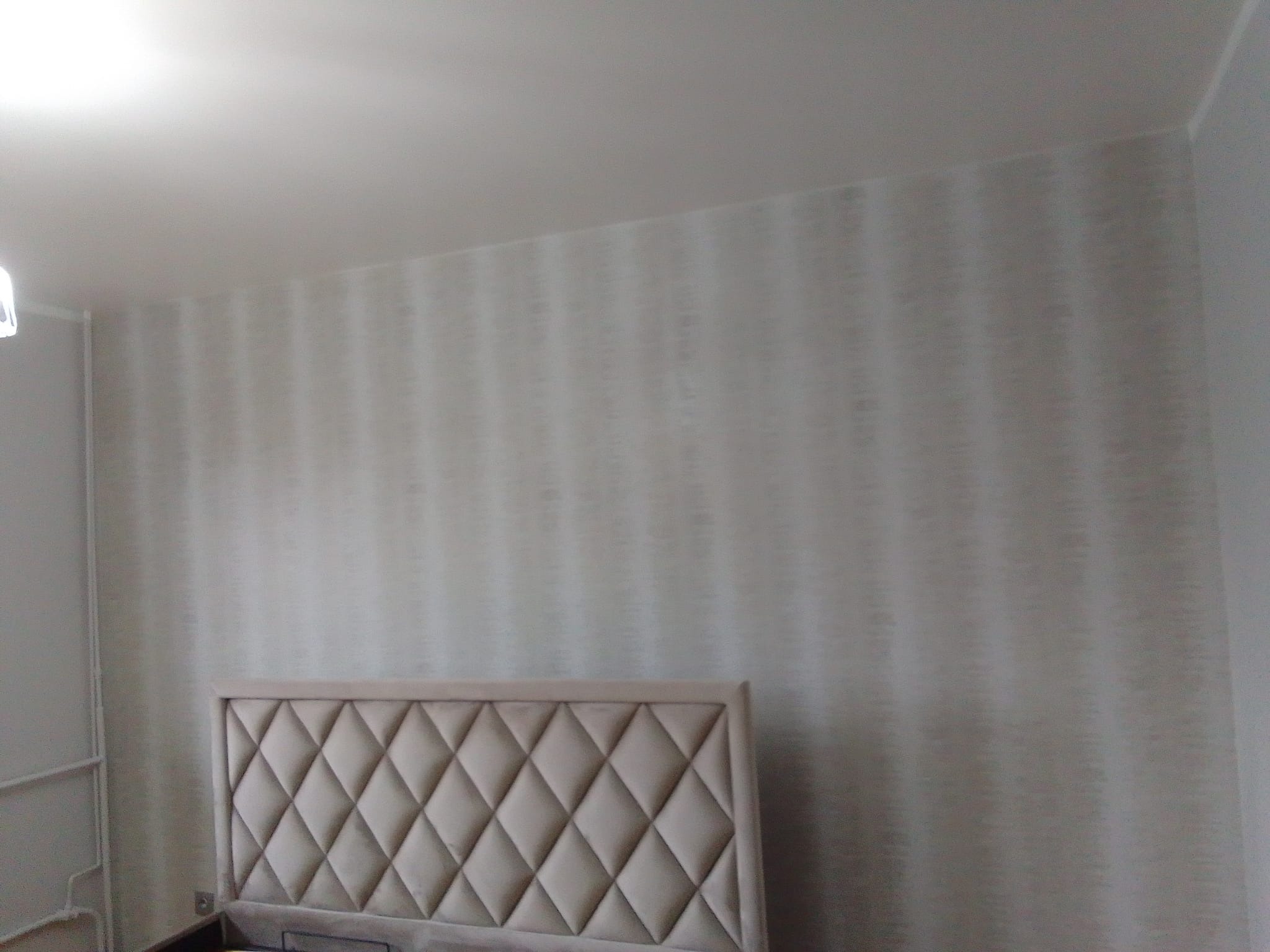 Ściana w sypialni pokryta tapetą w delikatne, szare prążki, widoczny fragment wezgłowia łóżka z beżową tapicerką w romby, sufit pomalowany na biało.