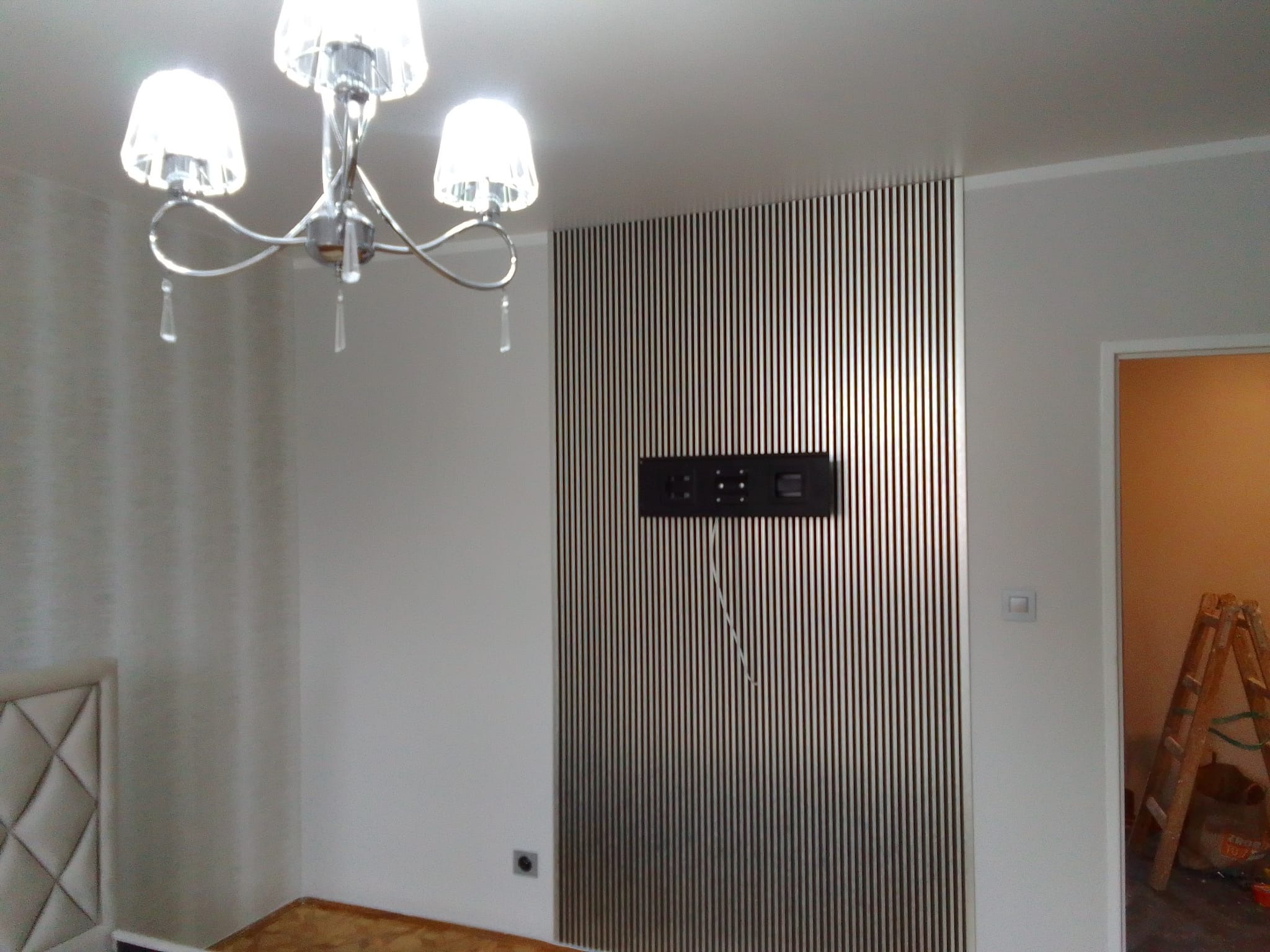 Nowoczesny pokój z panelami ściennymi w pionowe paski, wiszącym telewizorem i kryształowym żyrandolem; widoczny fragment łóżka i drabina oparta o ścianę przy drzwiach.