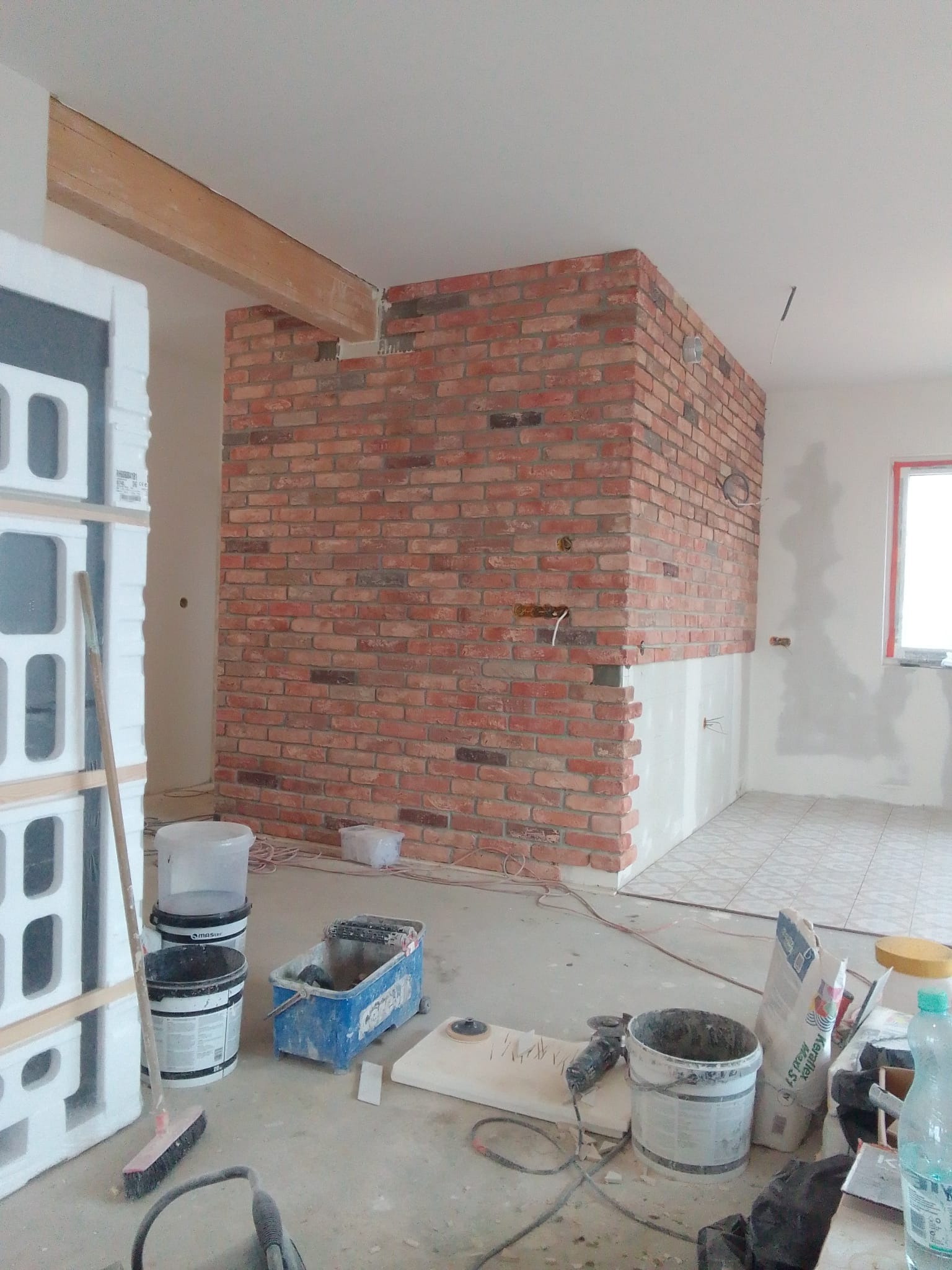 Wnętrze w trakcie prac wykończeniowych: fragment ściany wyłożony czerwoną cegłą, częściowo otynkowany dół ściany, widoczne puszki elektryczne, na podłodze narzędzia i materiały budowlane, w tle...