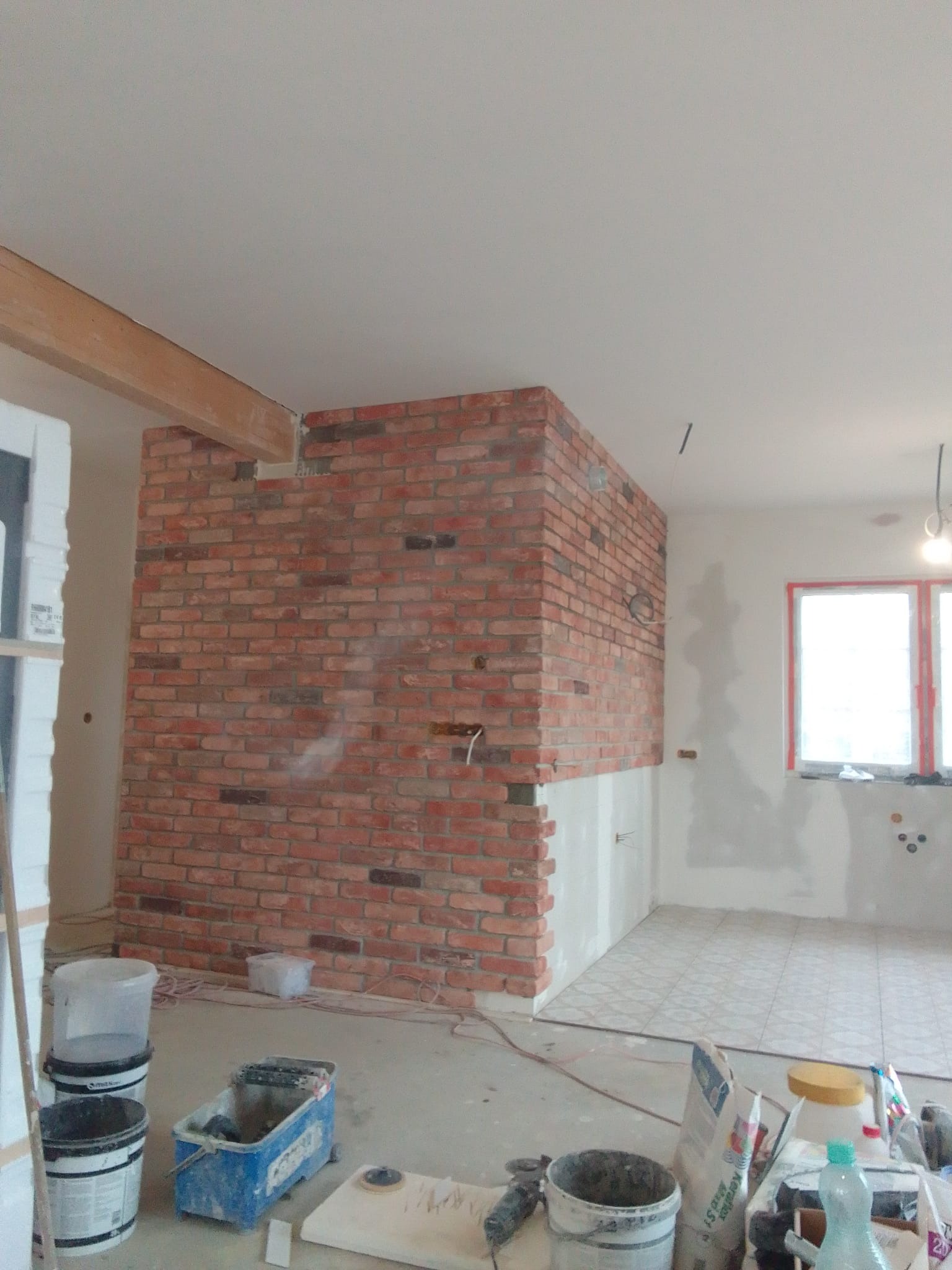 Ściana z czerwonej cegły dekoracyjnej w trakcie montażu, widoczne narzędzia i materiały budowlane, fragment okna w tle, surowy stan wykończenia wnętrza.